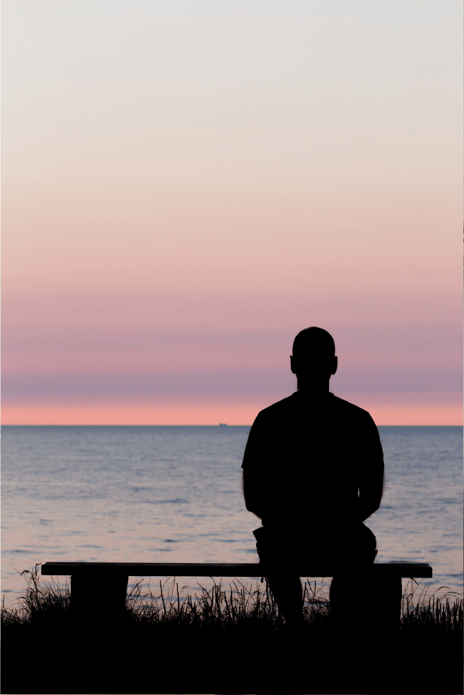 man's silhouette sitting on a bench looking at the ocean