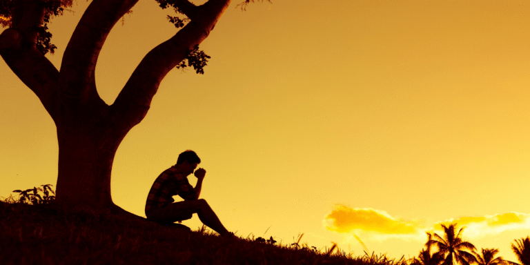 man on a hill under a tree with hands folded praying to God about his situation