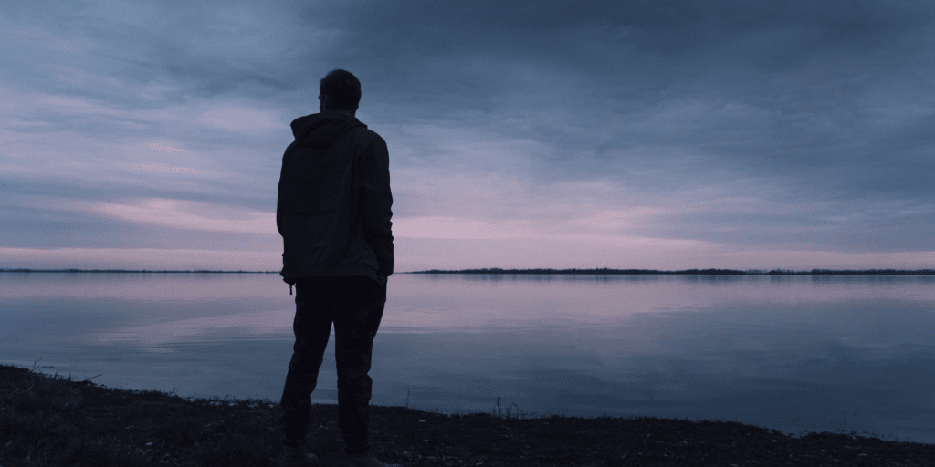 man standing outside by a lake and thinking while the sun goes down