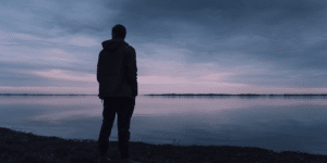 man standing outside by a lake and thinking while the sun goes down