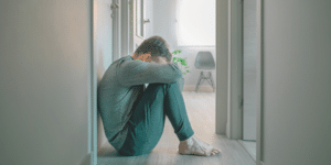 man hugging his knees with his head bowed down on the floor to symbolize frustration, anger, loneliness, or stress