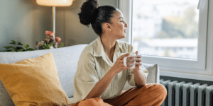 woman looking optimistically toward a window while drinking a beverage and sitting on her couch