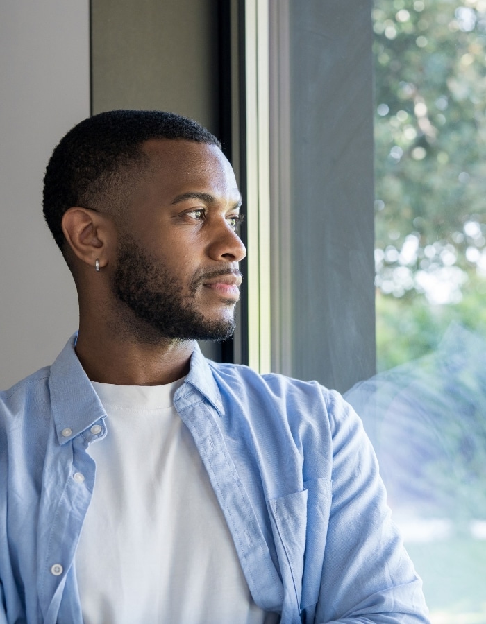 Thoughtful young Black man looking out a window, representing men's mental health counseling in Denver