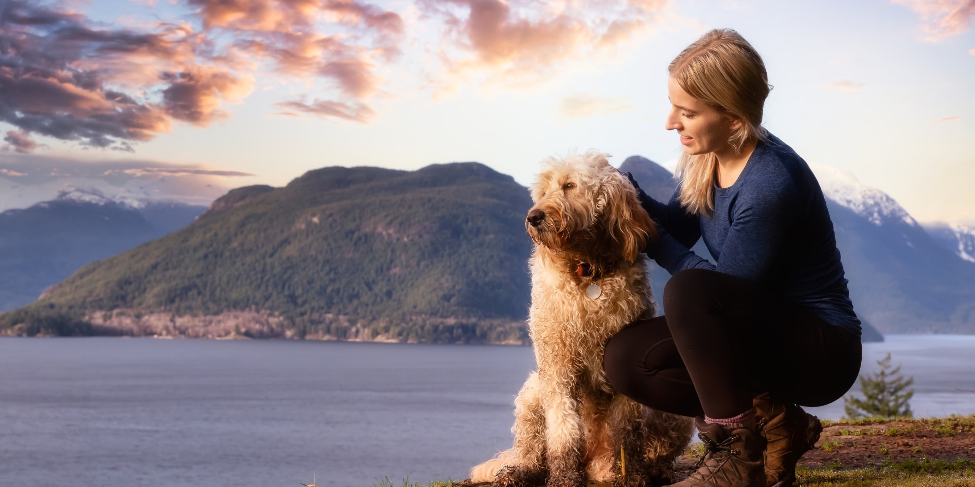 Woman kneeling with her dog overlooking mountains and a lake, honoring the grief of losing a pet