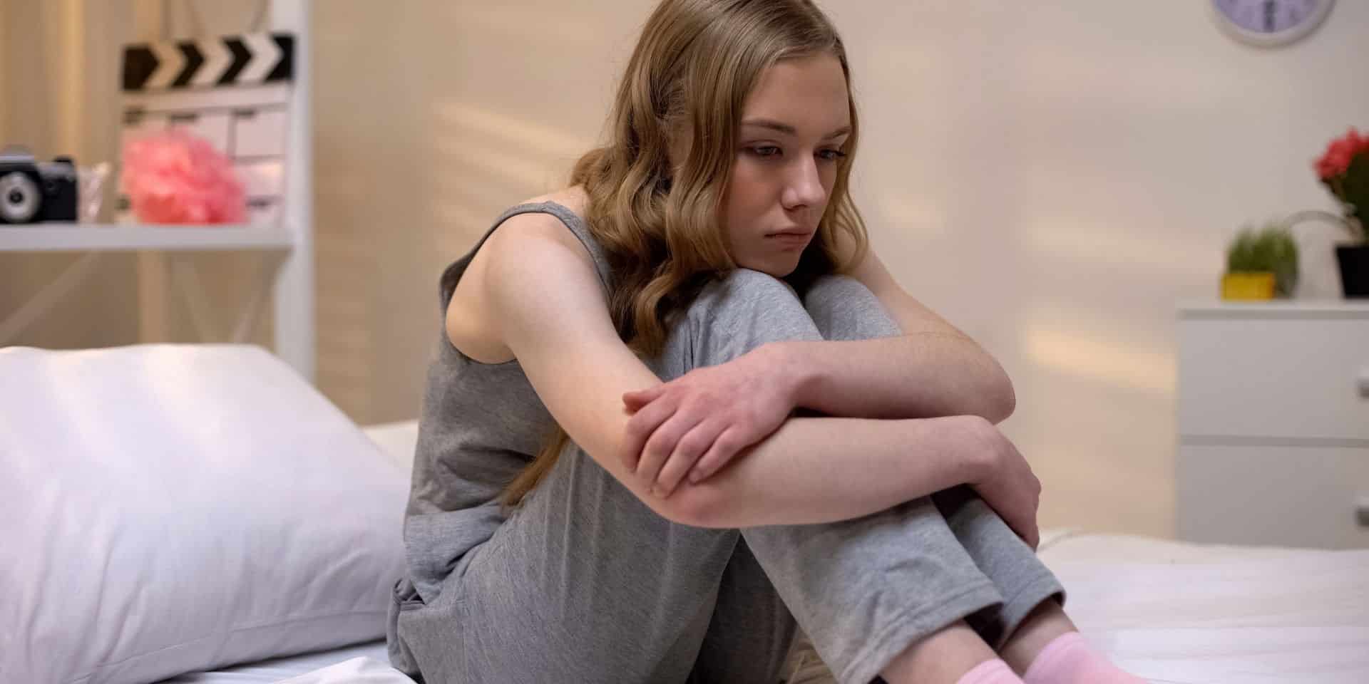 Teenage girl sitting on her bed hugging her knees, looking sad